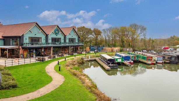 A riverside building with a docked boat, a grassy path, and a clear sky.