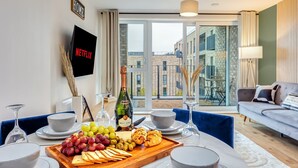 A modern living room with a dining area, a flat-screen TV, a bottle of champagne, and a view of the city through large windows.