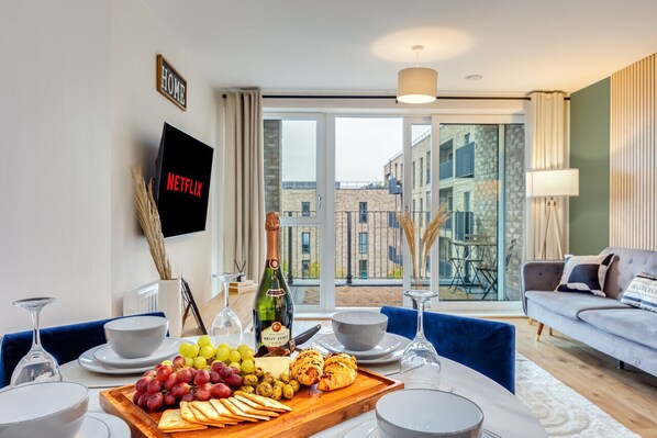 A modern living room with a dining area, a flat-screen TV, a bottle of champagne, and a view of the city through large windows.