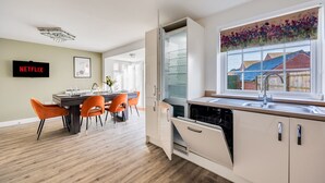 Una cocina moderna con lavavajillas integrado, un área de comedor con sillas color naranja y un ventanal con vistas a un jardín floral.