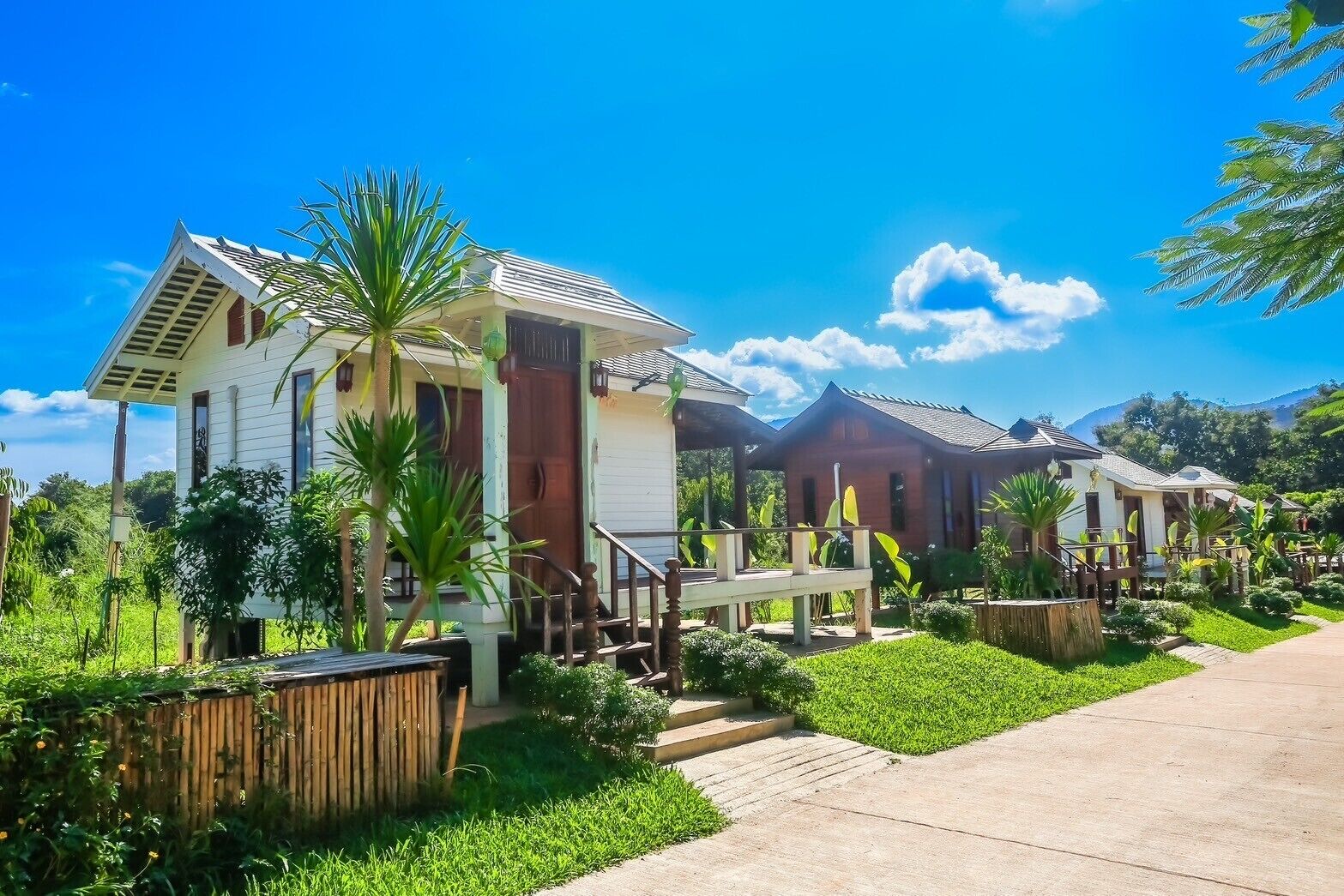 A row of single-story houses with a central walkway, surrounded by lush greenery and a clear blue sky.