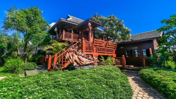 Ein traditionelles Holzhaus mit einer Treppe zu einem Balkon, umgeben von üppiger Vegetation und einer dekorativen Steinskulptur.