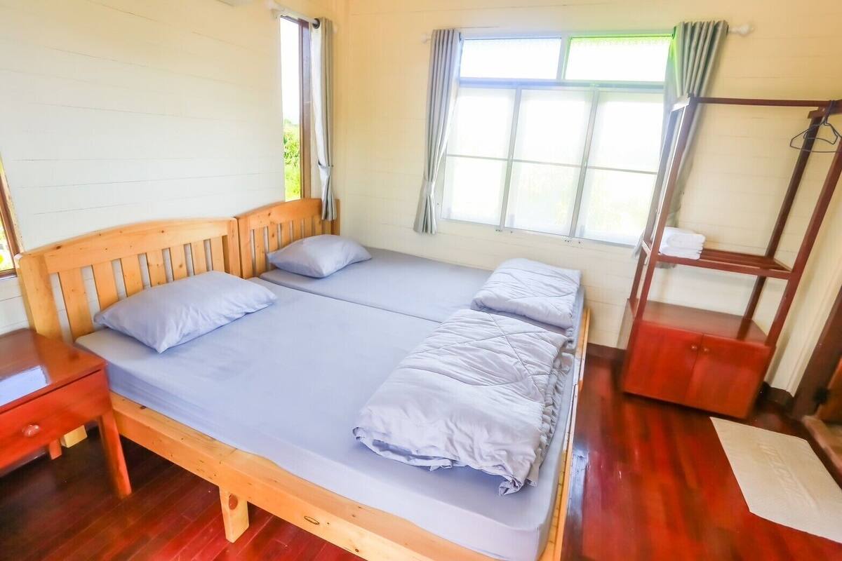 A room with a wooden bed, two pillows, a nightstand, a window with curtains, and a wooden shelf with towels.