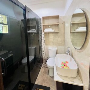 A bathroom with a glass shower enclosure, two toilets, a sink with a basin, and a mirror. - Top Ranks Hotel Galaxy (Yenagoa)