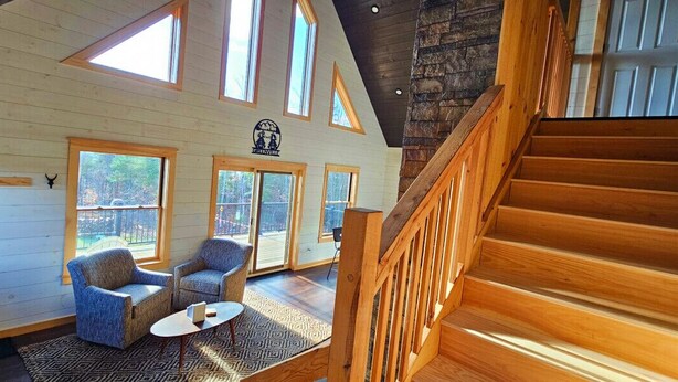 Une pièce spacieuse avec un escalier en bois, de grandes fenêtres et un coin salon confortable comprenant deux fauteuils bleus et une petite table.