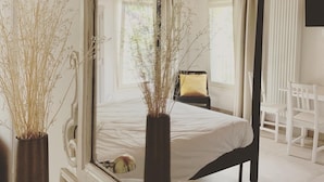 Una cama con dosel, un espejo encima, una silla blanca y un florero con plantas secas.