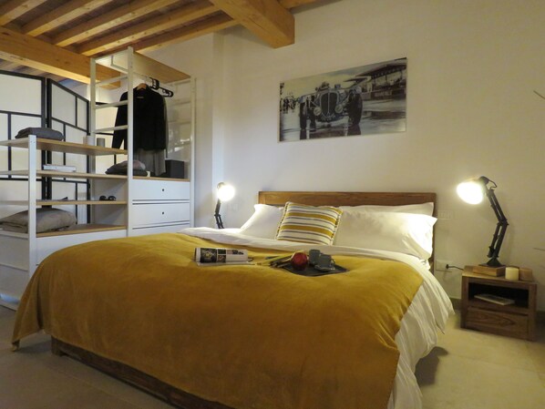A bedroom with a bed, bedside lamps, a nightstand, a shelf unit, and a wall-mounted picture.