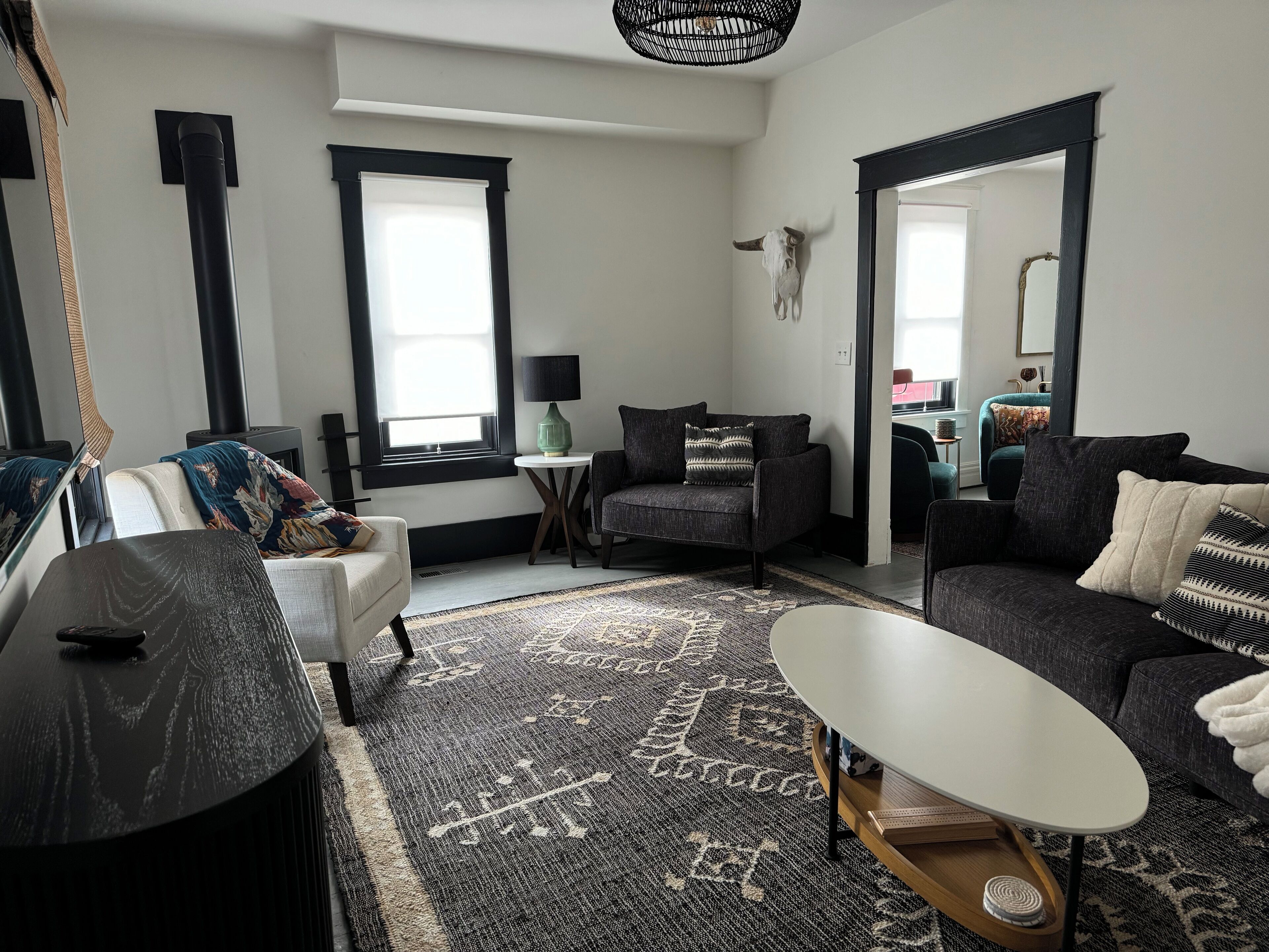 Una sala moderna con un sofá, un sillón y una mesa de centro. Hay una ventana con persianas, una lámpara y una calavera decorativa en la pared.
