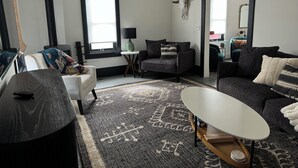 Ein modernes Wohnzimmer mit einer Couch, einem Sessel und einem Couchtisch. Es gibt ein Fenster mit Jalousien, eine Lampe und einen dekorativen Totenkopf an der Wand.