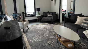 Una sala moderna con un sofá, un sillón y una mesa de centro. Hay una ventana con persianas, una lámpara y una calavera decorativa en la pared.