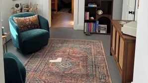 Ein Zimmer mit einem salbeengrünem Sessel, einem gemusterten Teppich, einem Holzregal und einer weißen Tür.