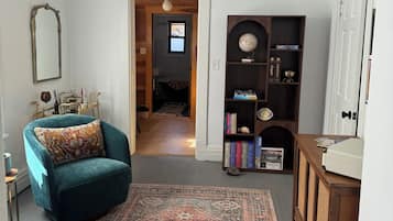 Una habitación con un sillón azul verdoso, un tapete estampado, una estantería de madera y una puerta blanca.