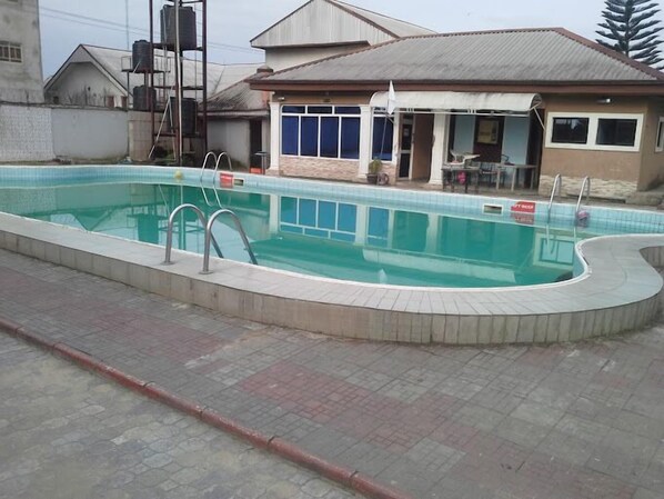 A swimming pool with a tiled deck, a covered seating area, and a building in the background.