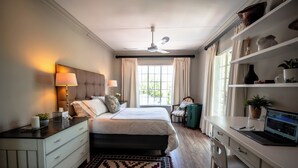 A bedroom with a bed, bedside tables, a desk with a laptop, a chair, a window with curtains, a ceiling fan, and a rug. - Crown Loft - Bright, Airy Stay with Balcony View (Sandton)