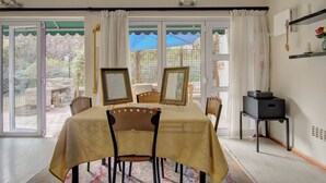 Un coin repas avec une table dressée pour quatre personnes, un haut-parleur noir posé sur un support et une vue sur une terrasse extérieure, visible à travers de grandes portes vitrées.
