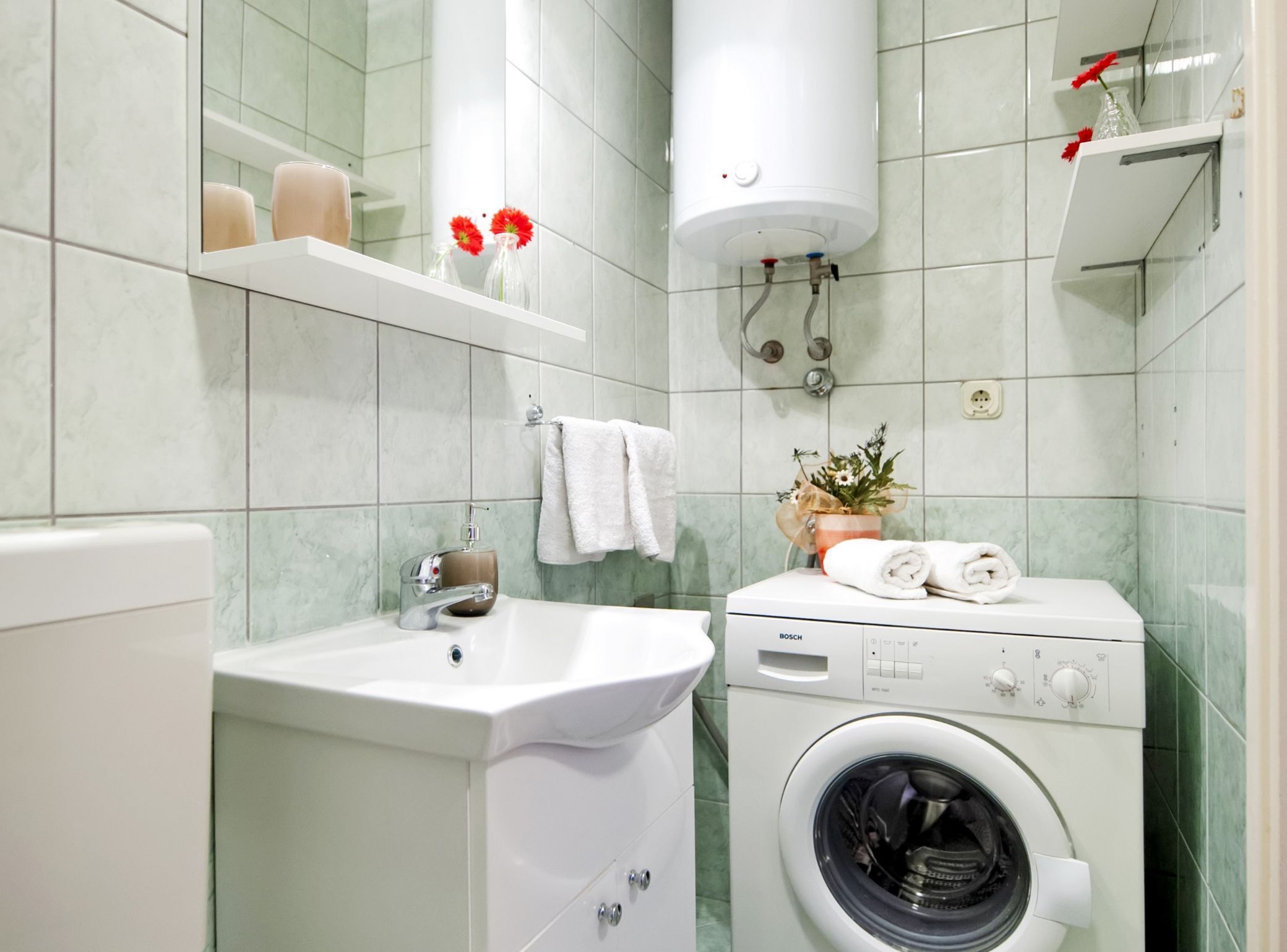 A modern bathroom with a sink, a Bosch washing machine, a towel rack, and a shelf with toiletries.
