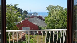 Une vue depuis un balcon doté d’une balustrade, donnant sur une zone côtière avec des bâtiments, des arbres et un ciel bleu dégagé.