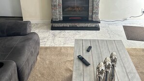 A living room with a stone fireplace, a flat-screen TV mounted above it, a dark leather sofa, and a coffee table with remote controls and decorative figurines. - Workers Heaven (Daleville)