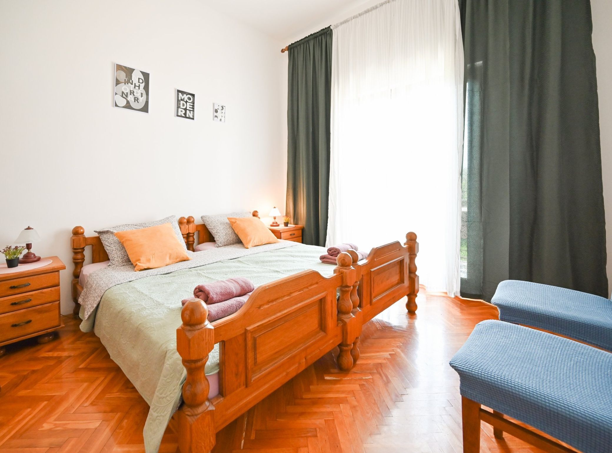 A bedroom with a wooden bed, two bedside tables, a nightstand, a blue chair, and a window with curtains.