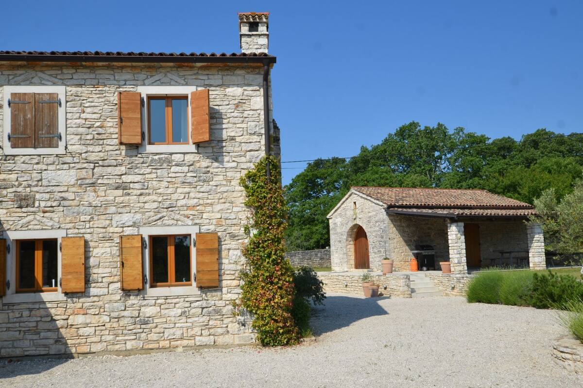 A stone house with wooden shutters, a gravel driveway, and a small attached structure with a tiled roof.