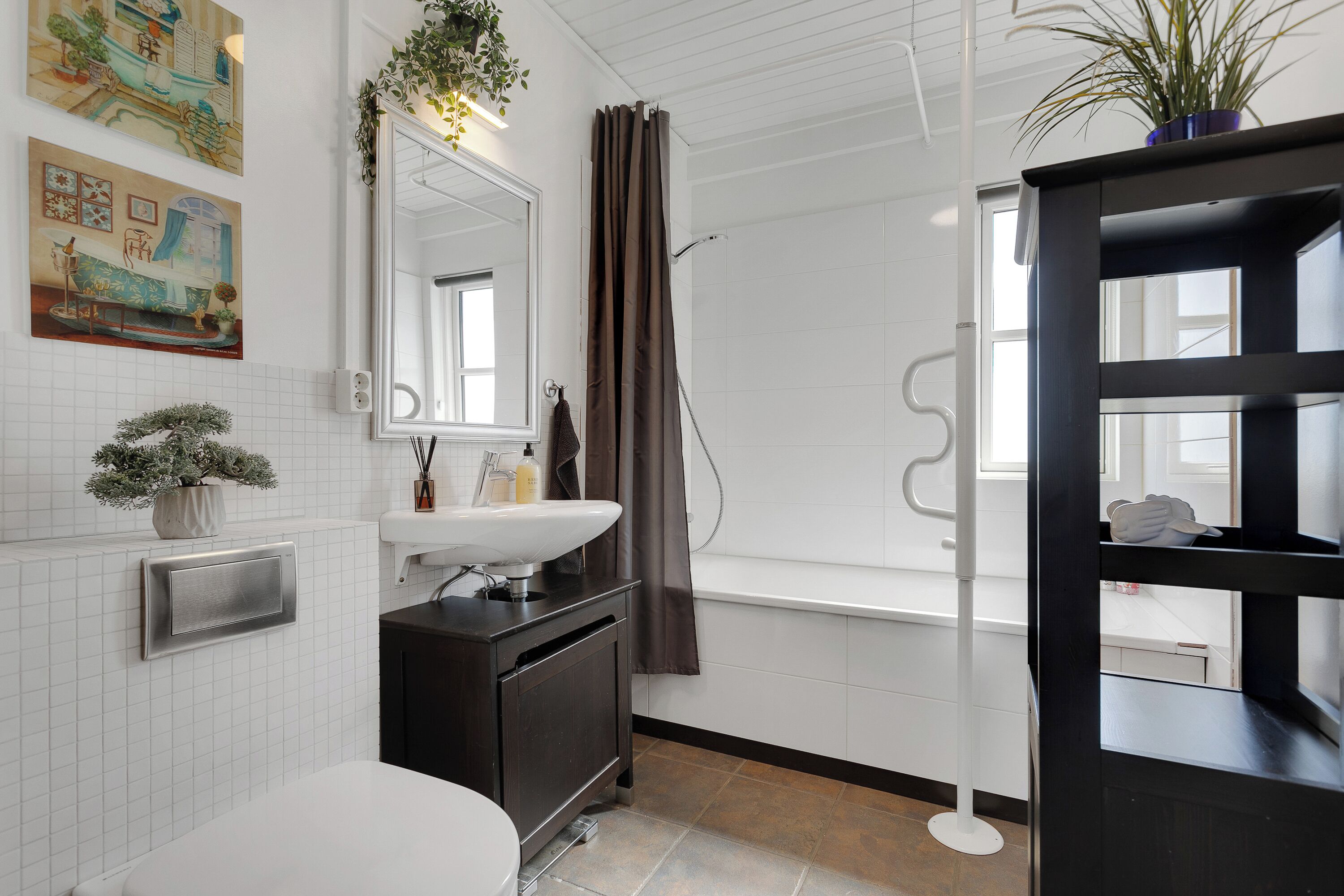 Un baño con lavamanos blanco, mueble oscuro y ventana con cortina de ducha.