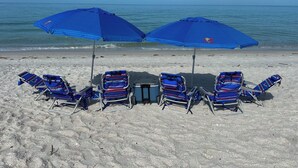 Een strand met zes blauwe ligstoelen en twee blauwe parasols in een rij.