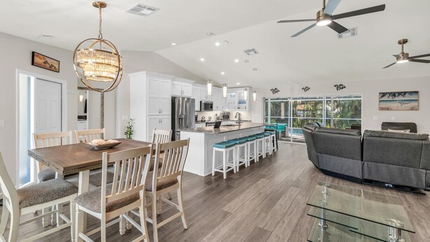 Een moderne keuken met witte kasten, een centraal eiland met barkrukken, een eethoek met een houten tafel en stoelen, en een woongedeelte met een grijze bank en glazen salontafel.