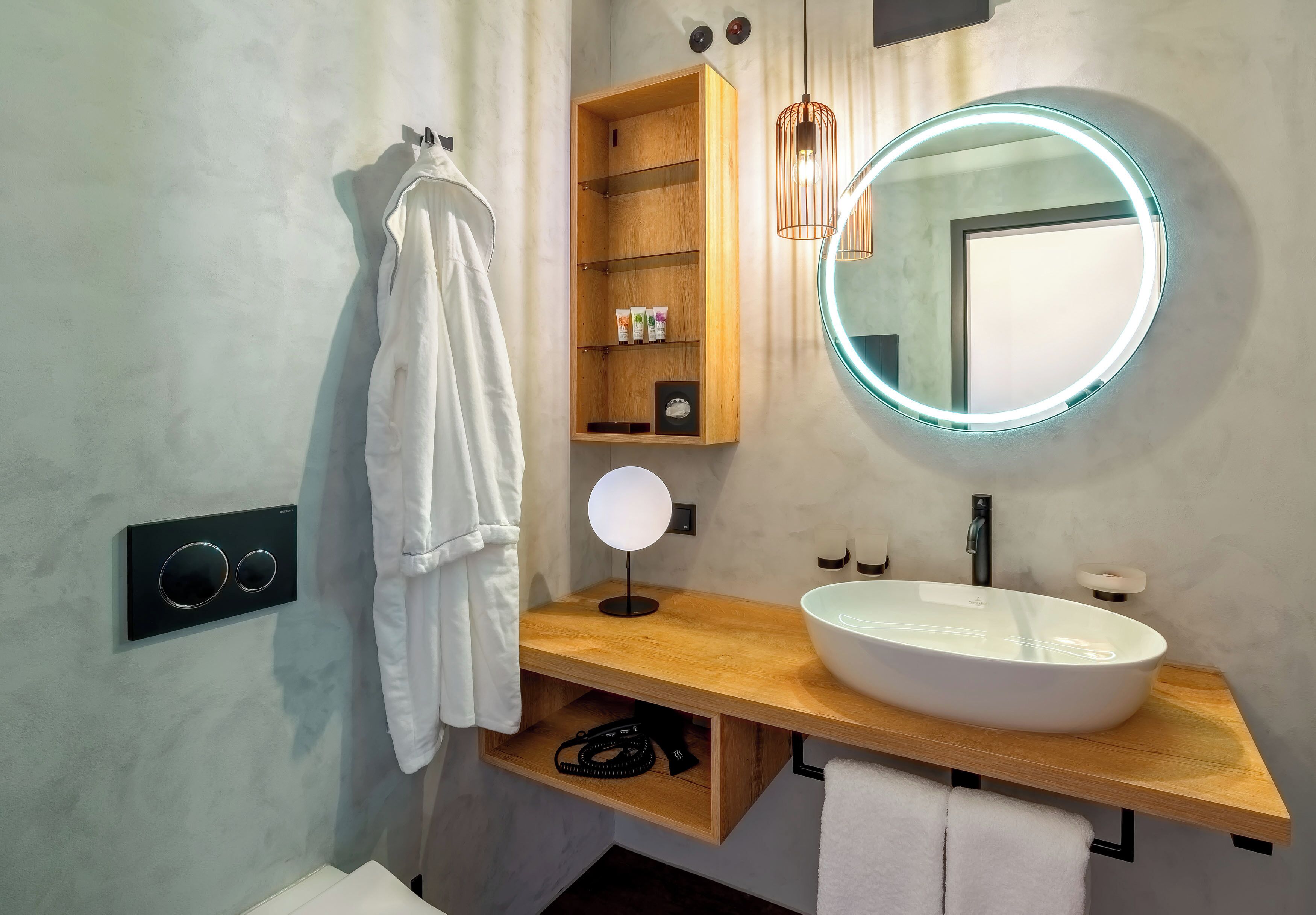 Een moderne badkamer met een houten wastafelmeubel, een ronde spiegel met verlichting, een witte wasbak en een handdoekhouder.