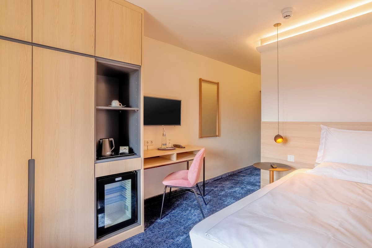 A hotel room with a bed, a desk with a chair, a television, a minibar, and a mirror.