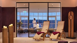 Wellnessbereich mit zwei Liegen, einem Blumentopf und Blick auf eine Person im Bademantel draußen.