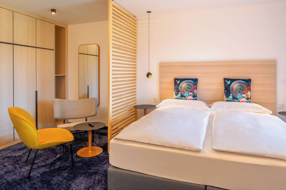 A hotel room with a bed, a chair, a small table, a mirror, and artwork on the headboard.