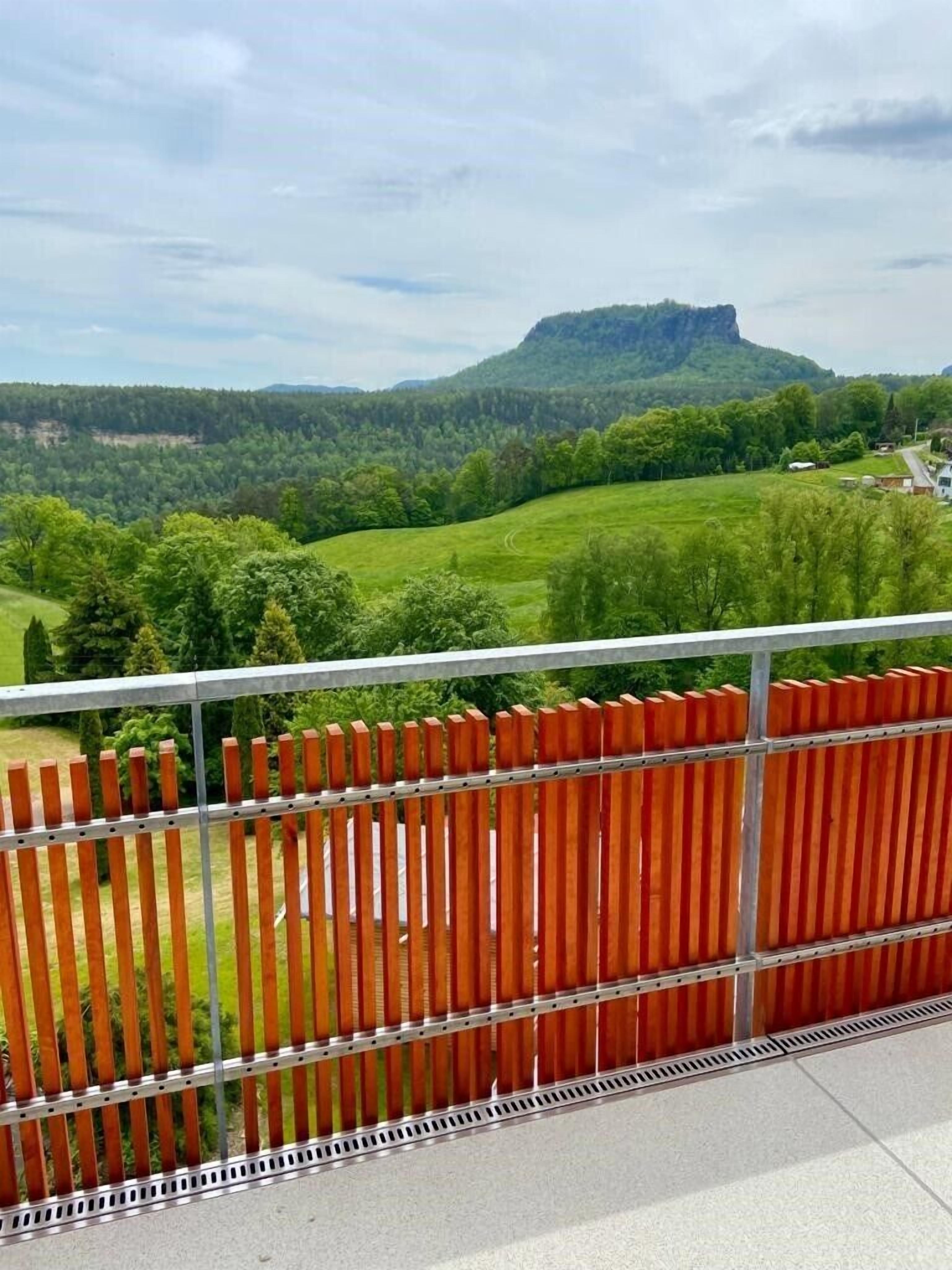 A balcony with a metal railing and a view of a green landscape with trees and a hill in the background.
