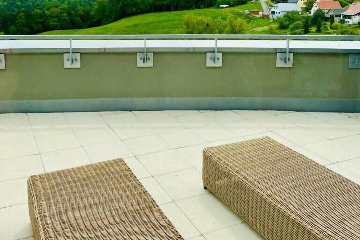 A rooftop terrace with two wicker lounge chairs, a view of a green landscape with a hill and houses in the background.