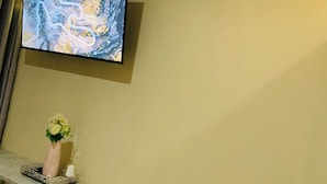 A room with a flat-screen TV mounted on the wall, an air conditioning unit, a decorative vase with flowers on a dresser, and LED strip lights along the ceiling.