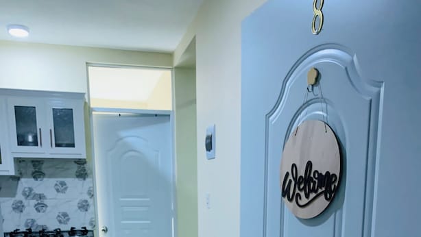 A room with a dark blue sofa, a white door with a 'Welcome' sign, and a kitchen in the background.