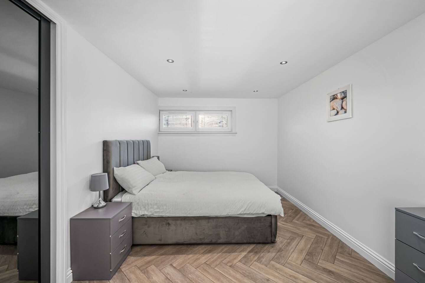 Une chambre à coucher avec un lit, une table de chevet, une commode, une fenêtre avec des stores et un tableau au mur.