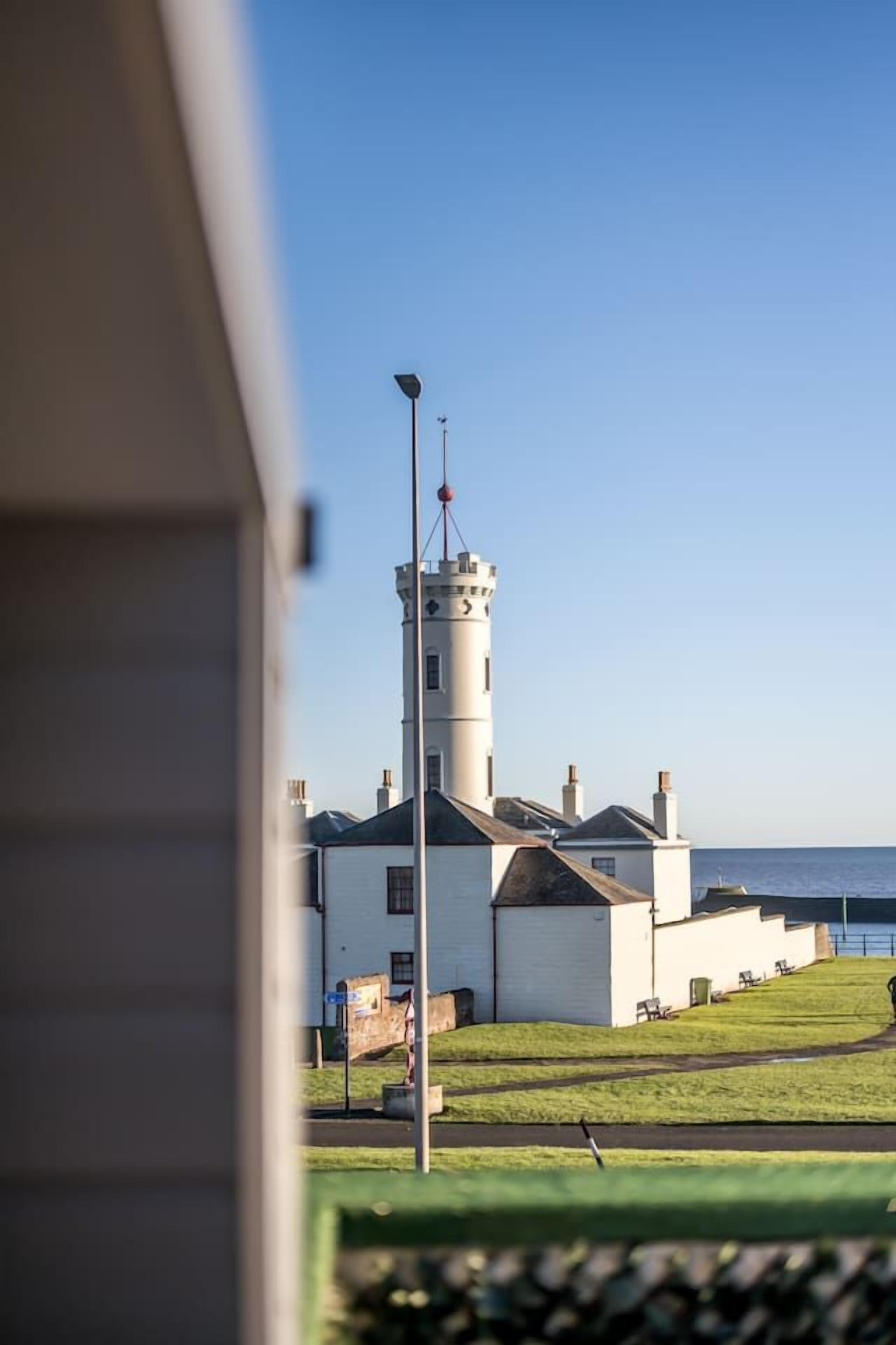 Un phare à l’extérieur blanc et doté d’une haute tour, situé dans une zone herbeuse offrant une vue dégagée sur la mer.