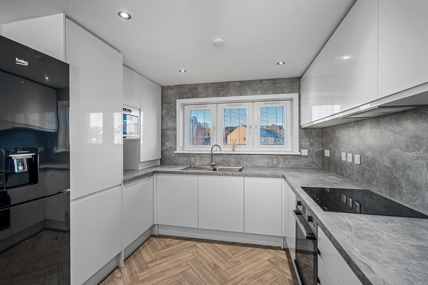 Een moderne keuken met witte kasten, een visgraat parketvloer, een ingebouwde oven en een raam met jaloezieën.