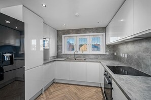 A modern kitchen with white cabinets, a herringbone wood floor, a built-in oven, and a window with blinds. - Sea Views Luxury Apartment Free Parking - Coast Ap (Arbroath)