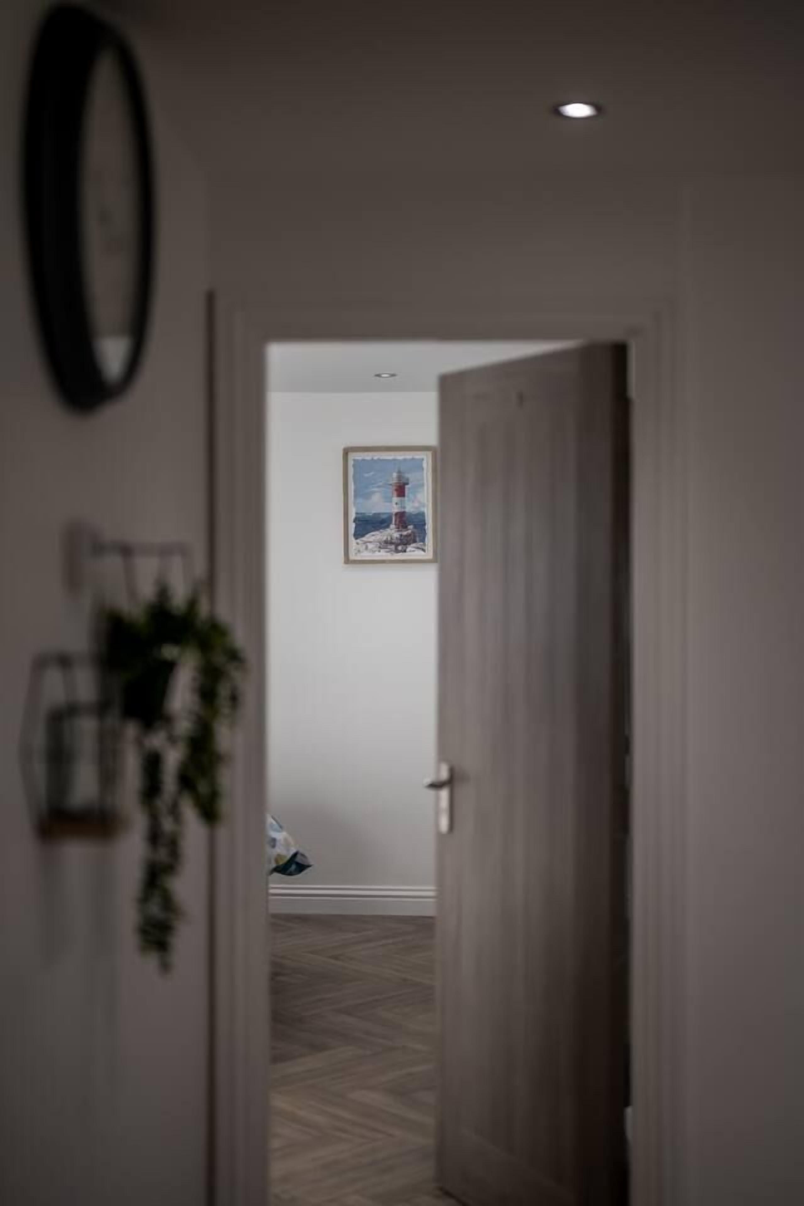 Um corredor com uma porta de madeira, um quadro na parede e uma planta no batente da porta.