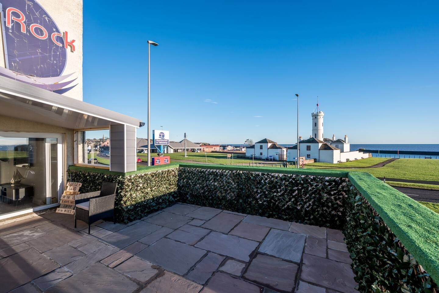 Een hotel aan de kust met een terras, uitzicht op zee en een vuurtoren in de verte.