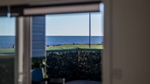 A view through a glass door to an outdoor patio area with a chair and a small table, overlooking a grassy area and a body of water. - Luxury Apartment with Sea Views Balcony (Arbroath)