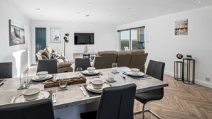 A modern dining area with a large table set for six, a sofa, and a flat-screen TV in the background. - Luxury Apartment with Sea Views Balcony (Arbroath)