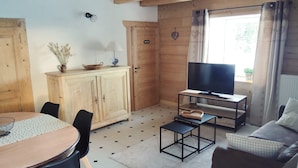Een gezellige kamer met houten plafond, een televisie op een standaard, een eettafel met stoelen en een bank.