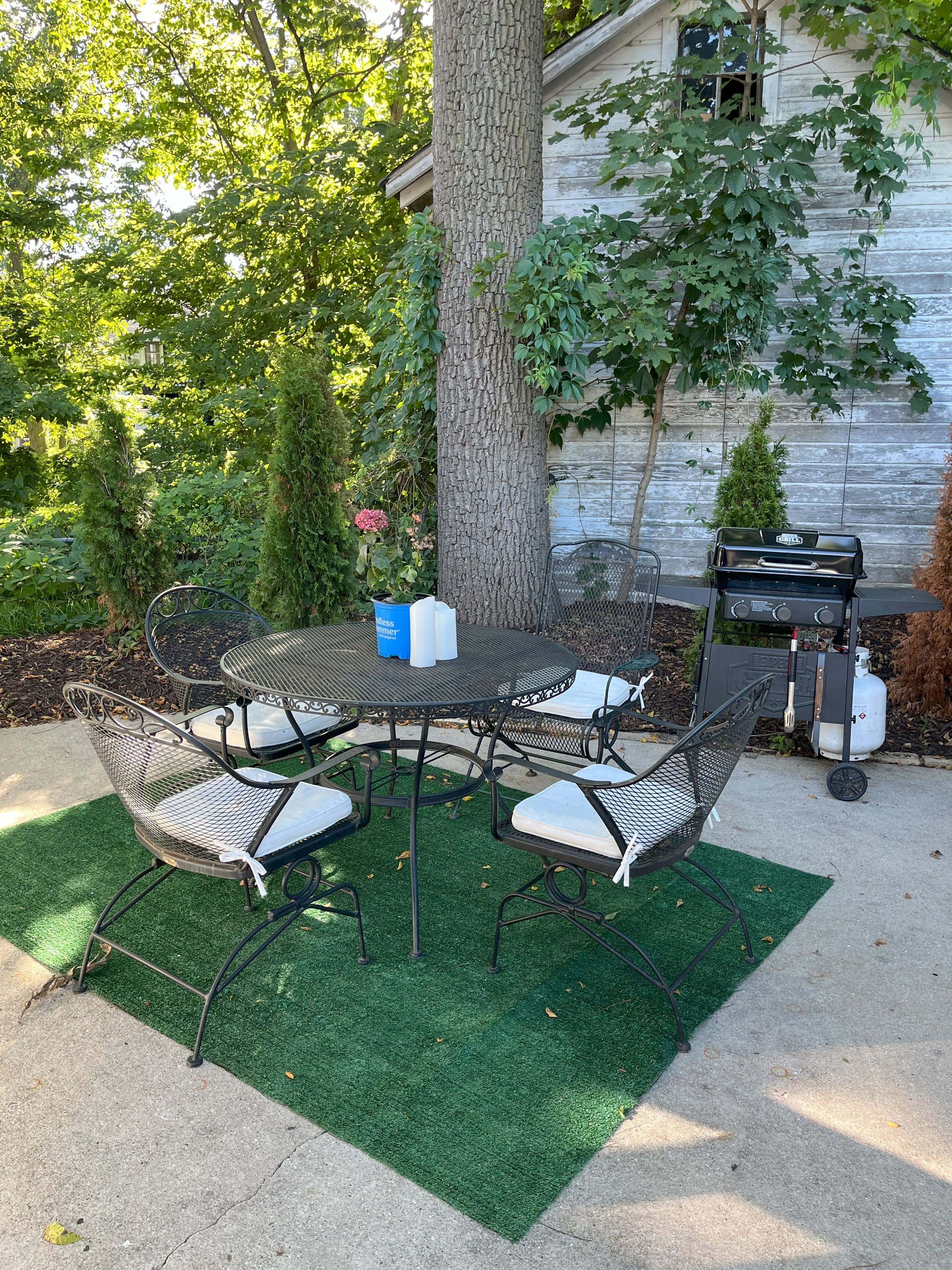 A round metal table with four chairs, surrounded by artificial green turf, with a tree and a grill in the background.