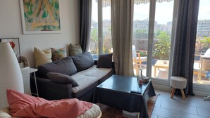 A living room with a sofa, a small table, a chair, and a view of the city through large windows. - Beautiful, quiet, bright artist's apartment.  South-facing terrace, air conditioning. (Marseille)