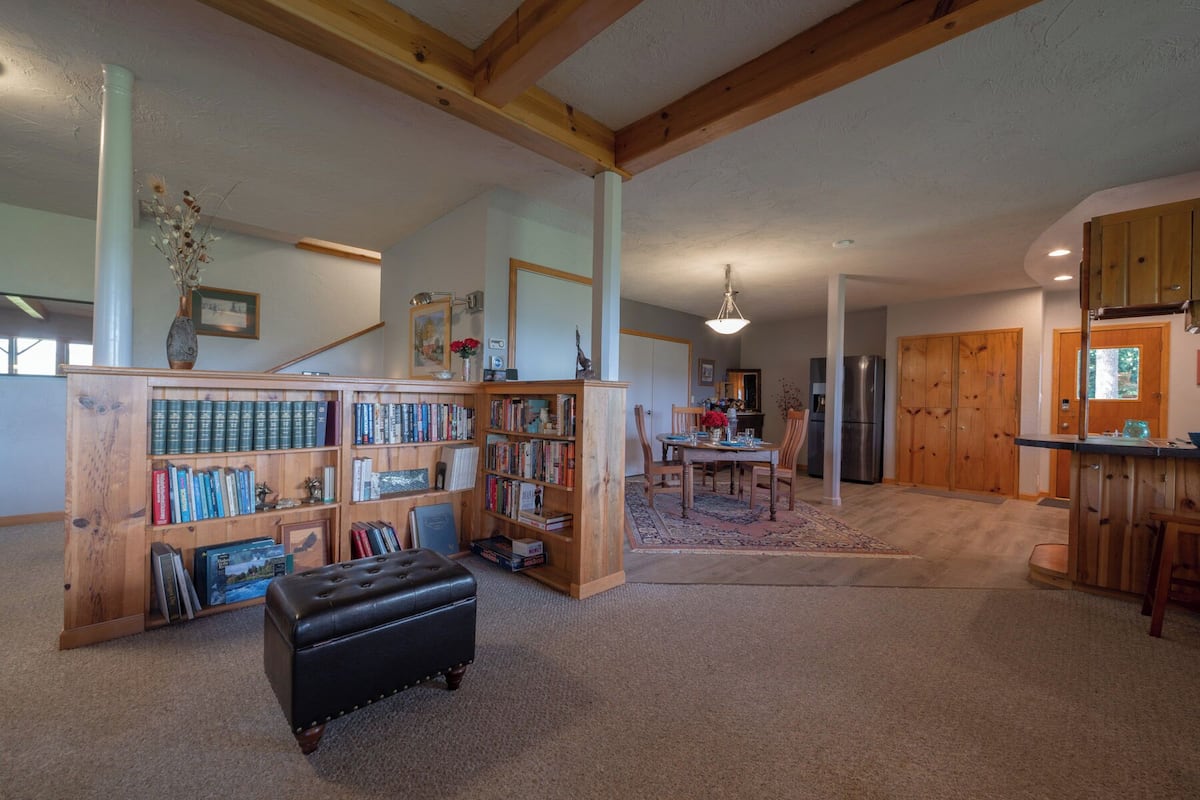 A spacious living room with a large wooden bookshelf, a dining area with a table and chairs, and a kitchen area with wooden cabinets and a refrigerator.