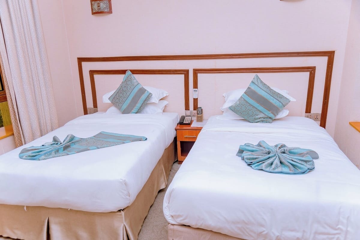 Two twin beds with white linens and blue towels, a wooden headboard, a bedside table with a phone, and a wall-mounted light switch.
