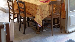 A dining area with a table set for four, surrounded by wooden chairs. There is a window with a view of greenery outside, and a door to the right. - Harvesters' house (Aigues-Vives)