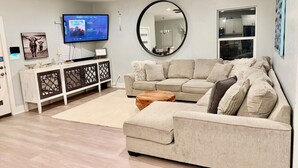 Ett vardagsrum med en beige soffa, ett soffbord, en tv på ett tv-bord, en takfläkt och en stor spegel.